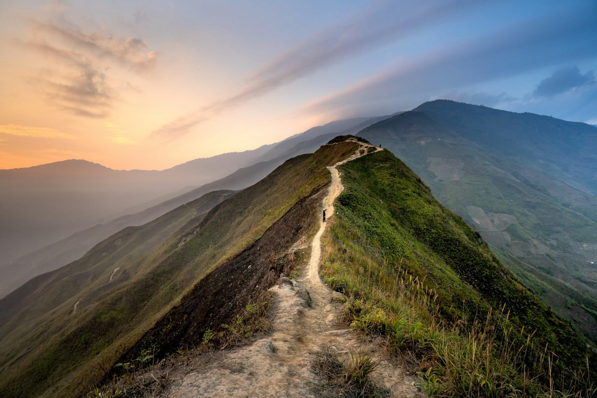 A winding mountain ridgeline trail with a lone hiker beneath a golden and blue sky